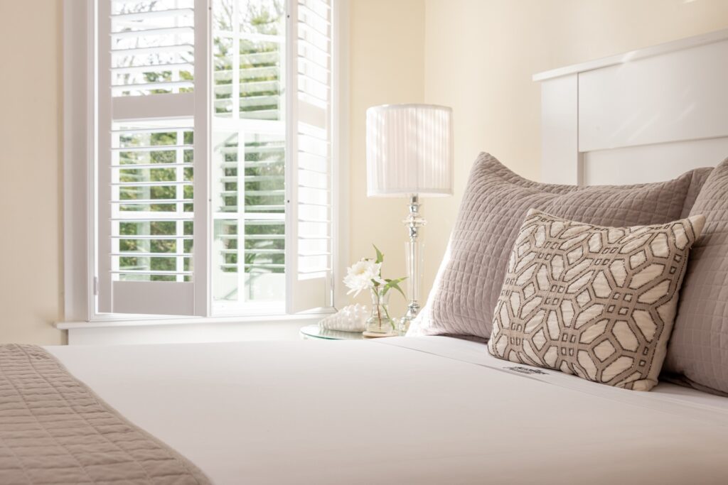 A guest room at the best bed and breakfast on Cape Cod, featuring a plush bed with throw pillows and a lamp on a nightstand beside a bright window filtering natural light.